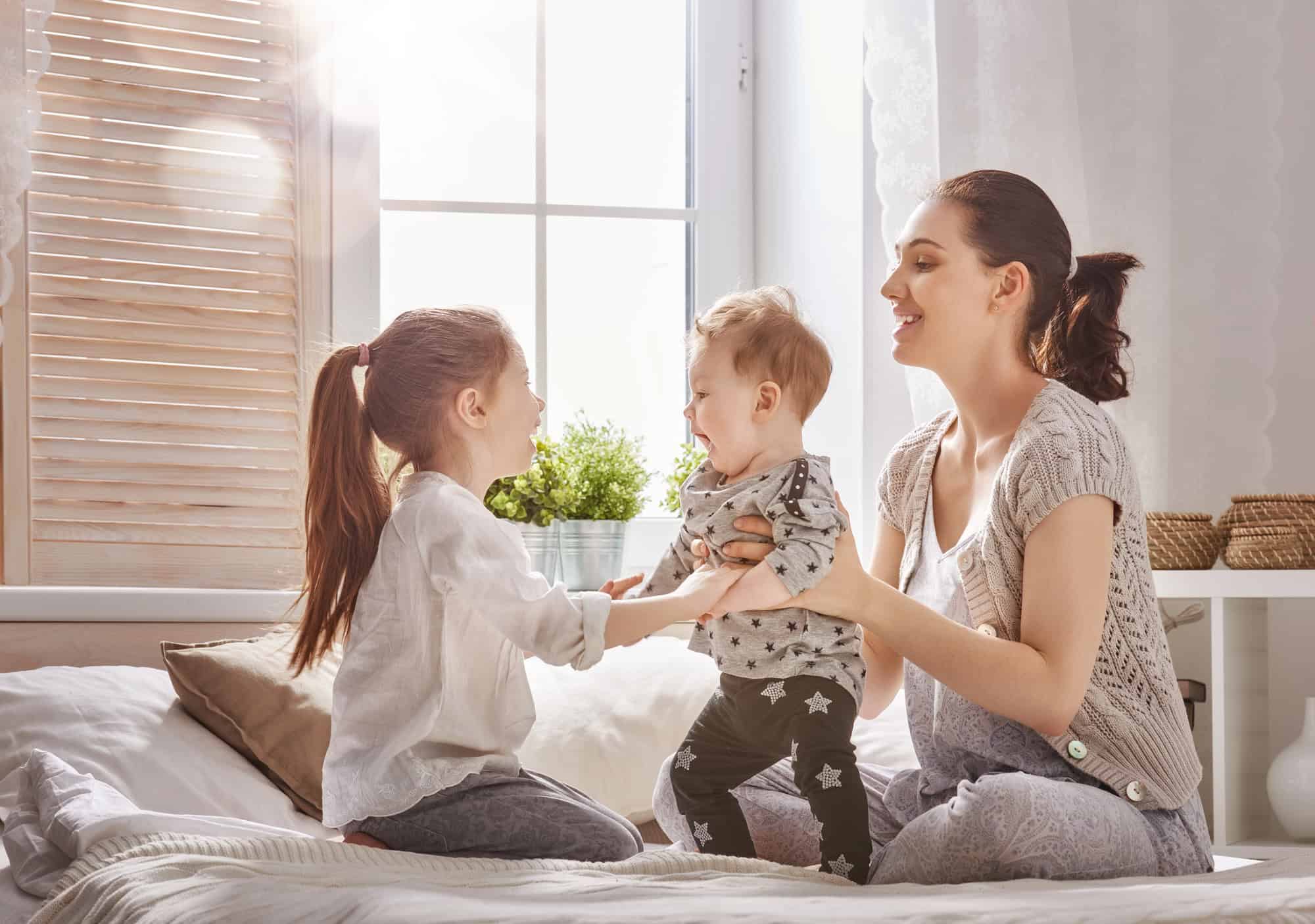 mom playing with her daughter and baby on the bed smiling and having a good time